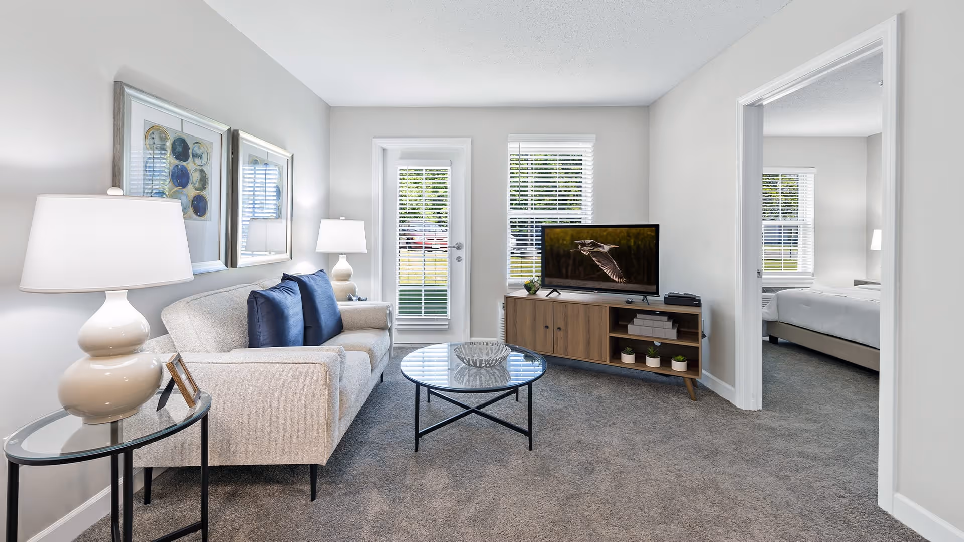A bright living room with a beige sofa adorned with blue pillows, a round glass coffee table, and a wooden TV stand holding a flat-screen television displaying a bird in flight. Two table lamps with white shades are placed on side tables beside the sofa. The room has light gray walls, carpeted flooring, and a door with window blinds leading outside. An adjacent bedroom with a bed and window is visible through an open doorway.