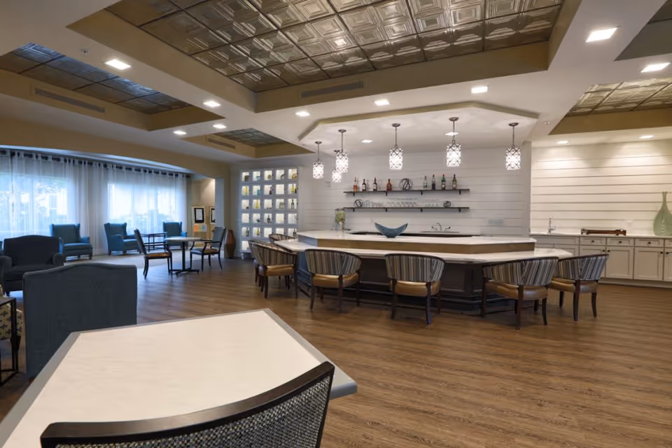 A spacious and modern common area with a large central bar counter surrounded by chairs, pendant lights hanging from the ceiling, shelves with bottles and decorative items on the wall, and several seating areas with armchairs and tables near large windows with sheer curtains.