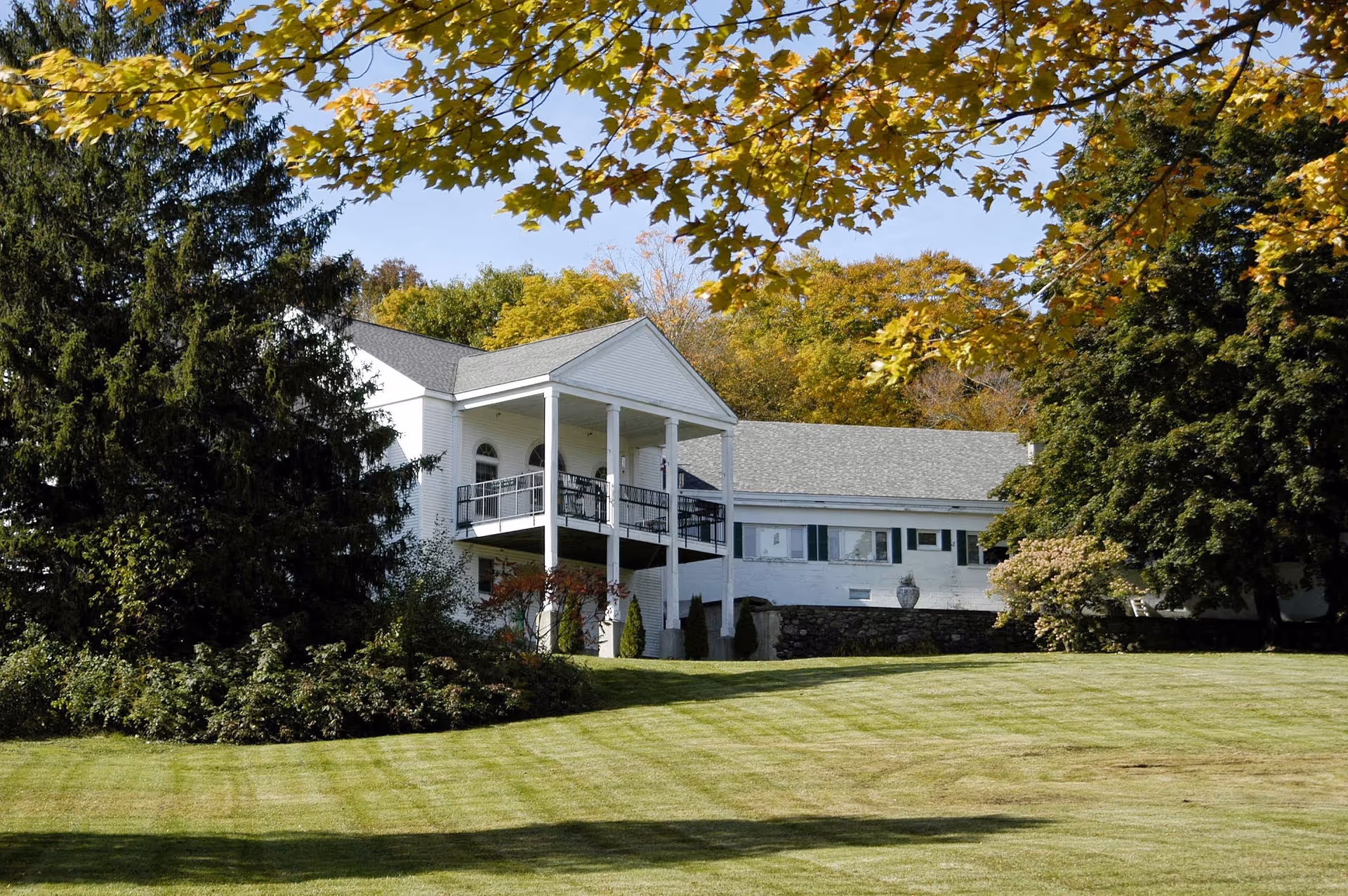 White two-story building with a columned balcony set behind a manicured lawn and trees with autumn foliage.
