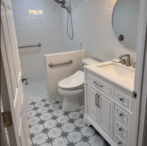 A clean and modern bathroom featuring a white toilet with a safety grab bar, a white vanity with a sink and faucet, a round mirror above the sink, and a walk-in shower with a handheld showerhead and grab bar. The floor has a patterned tile design in shades of gray and white, and the walls are covered with white subway tiles.