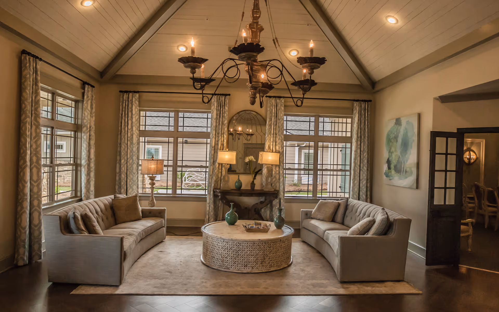 A cozy living room with two beige sofas facing each other across a round, ornate coffee table. The room features large windows with patterned curtains, a chandelier hanging from a vaulted ceiling, and a console table with lamps and decorative vases beneath a mirror. A painting is on one wall and a doorway leads to another room with chairs.