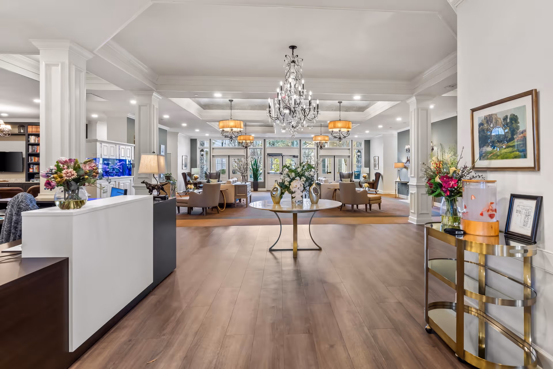 Spacious and elegant senior living facility common area with a chandelier, comfortable seating arrangements, a round table with floral centerpieces, and a reception desk on the left. The room features wood flooring, large windows, and decorative columns.