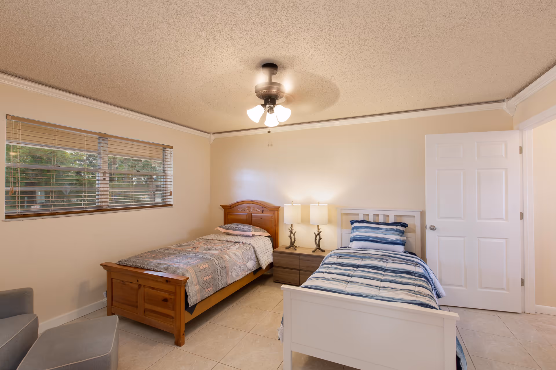 A bedroom with two single beds, one with a wooden frame and patterned bedding, the other with a white frame and blue striped bedding. Between the beds is a nightstand with two lamps. There is a window with blinds on the left wall and a ceiling fan with lights above. A gray armchair and ottoman are partially visible in the corner.