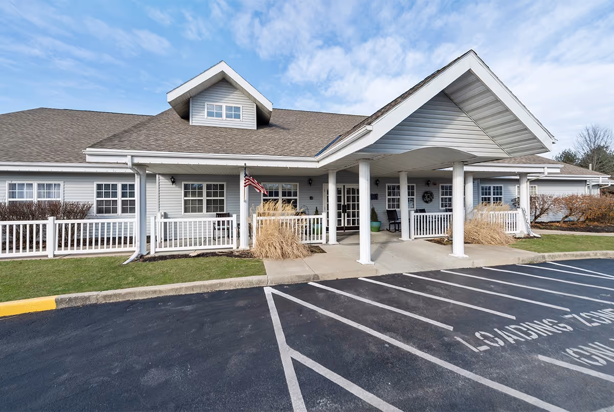 Front entrance of a single-story assisted living building with a covered porte-cochere, white railings, and adjacent parking/loading area.