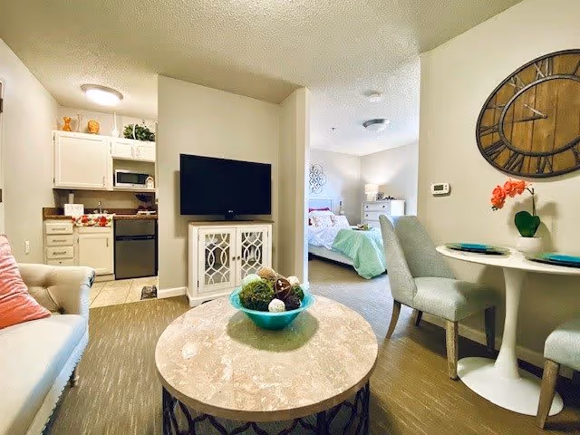A cozy senior living apartment interior featuring a small living room with a beige sofa and a round marble coffee table with a decorative bowl. A flat-screen TV is mounted on a white cabinet. To the left, there is a compact kitchen with white cabinets and appliances. To the right, a small dining area with a round white table and two upholstered chairs is visible, along with a large wall clock and a potted orchid. In the background, a bedroom with a bed, white dresser, and soft lighting can be seen.