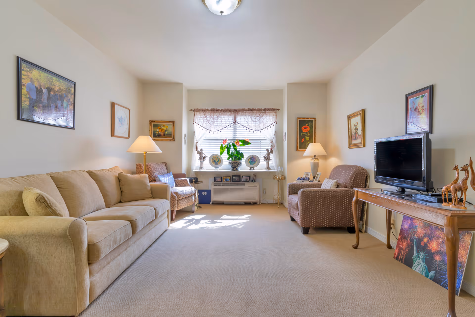 A cozy living room with a beige sofa on the left, two patterned armchairs on either side of a window with sheer curtains, and a wooden table on the right holding a flat-screen TV and decorative giraffe figurines. The room is softly lit by two table lamps and decorated with framed pictures on the walls.
