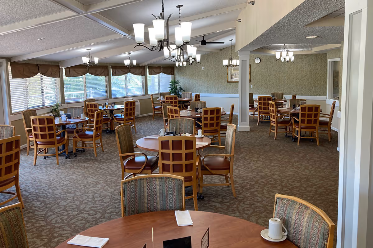 A spacious dining room with multiple round tables and wooden chairs arranged neatly. The room has large windows with blinds allowing natural light to enter, patterned carpet flooring, and several ceiling light fixtures providing warm lighting. The tables are set with napkins, cups, and condiments, creating a welcoming atmosphere.