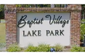 A brick sign with the text 'Baptist Village LAKE PARK' surrounded by grass and some greenery.