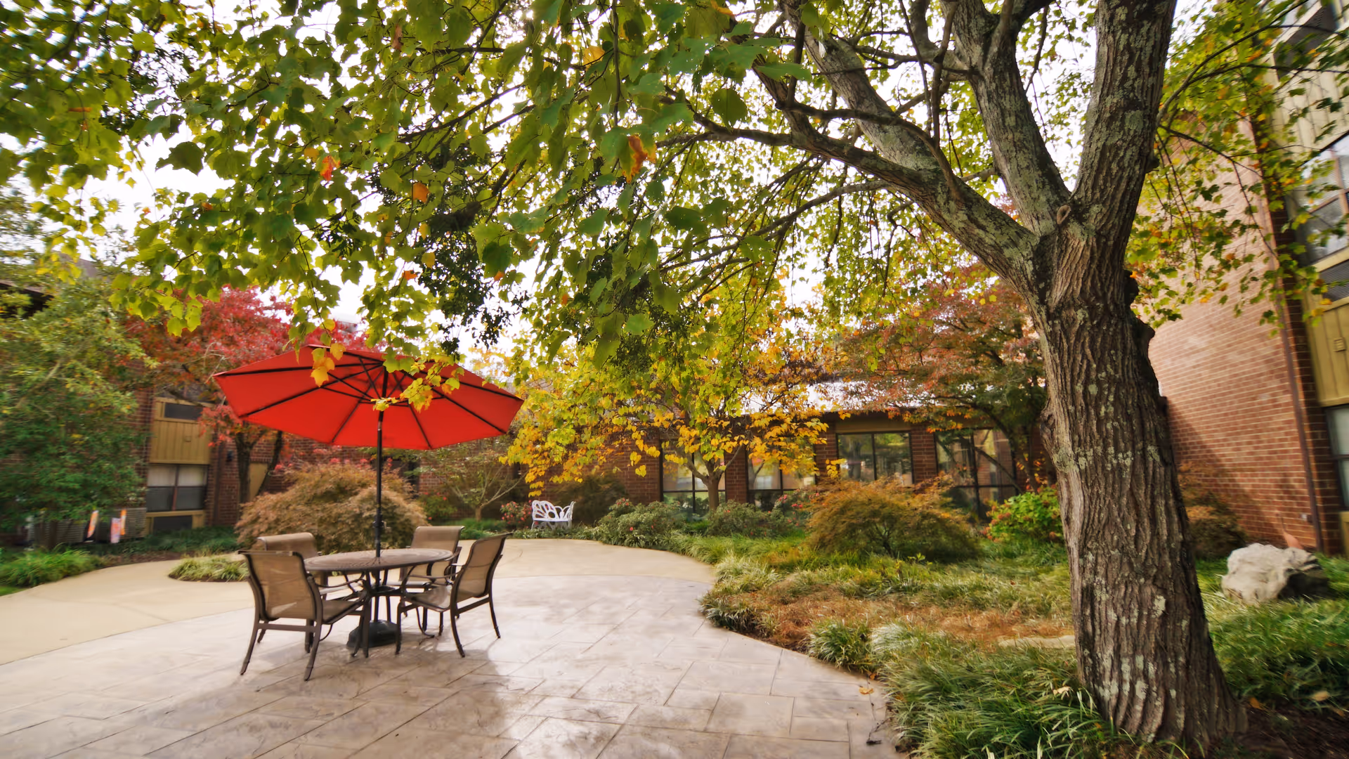 Outdoor patio area at a senior living community with a round table, four chairs, and a large red umbrella. The patio is surrounded by lush greenery, trees with autumn-colored leaves, and a brick building in the background.