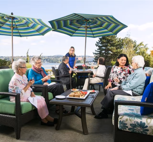 A group of elderly people and a caregiver sitting and chatting outdoors under large striped umbrellas. They are seated on cushioned patio furniture around a table with plates of food and drinks, with a scenic view of trees and water in the background.