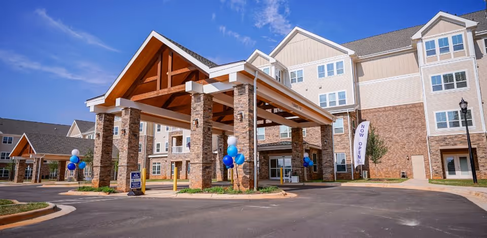 Exterior view of a senior living facility named Harmony at Brookberry Farm featuring a large covered entrance with stone pillars and wooden beams, decorated with blue and white balloons. The building is multi-story with beige and brick facade under a clear blue sky.