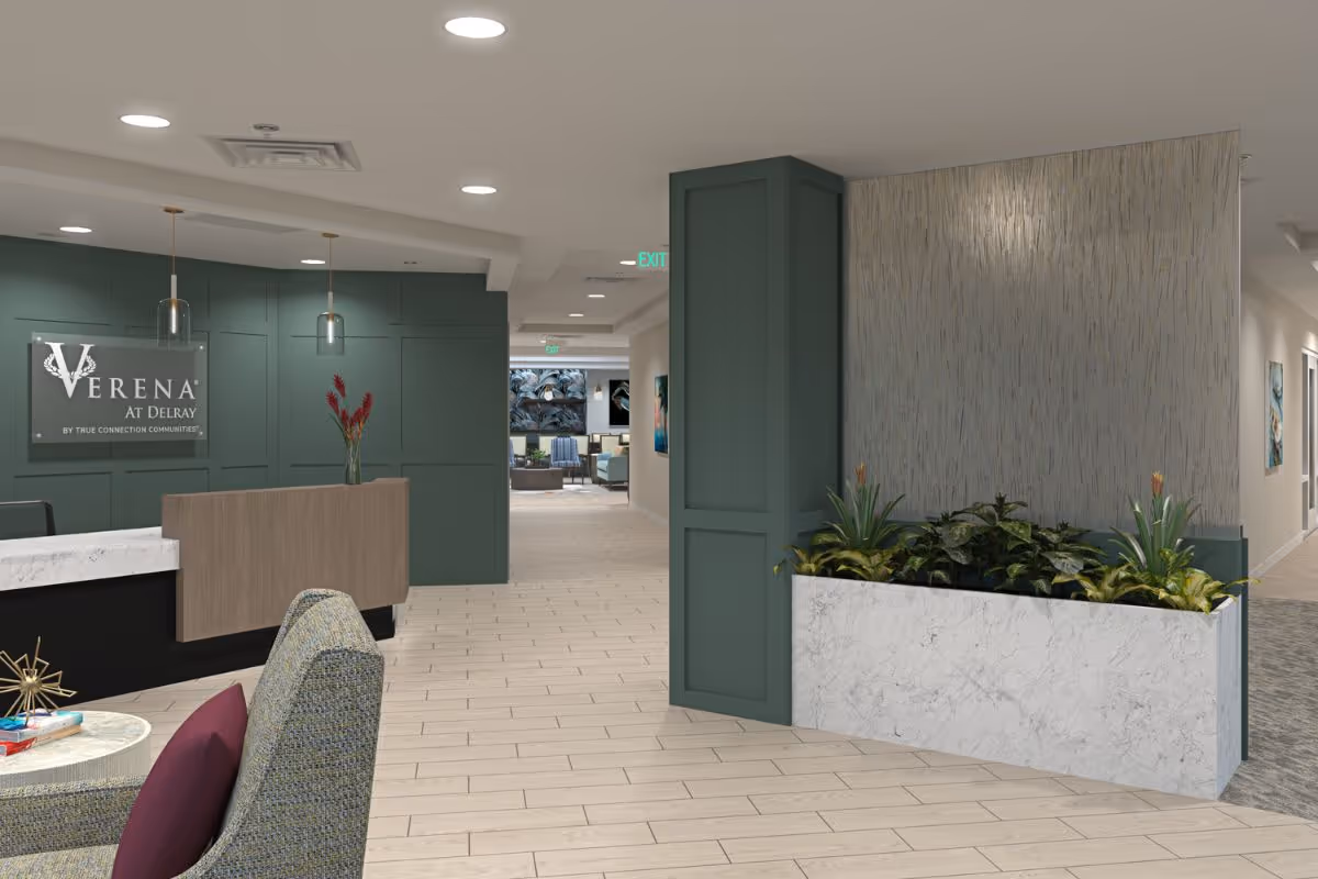 Interior view of the reception area at Verena at Delray featuring a modern front desk with a wooden and marble finish, green paneled walls, pendant lights, a planter with green plants, and a hallway leading to a lounge area with seating and artwork on the walls.