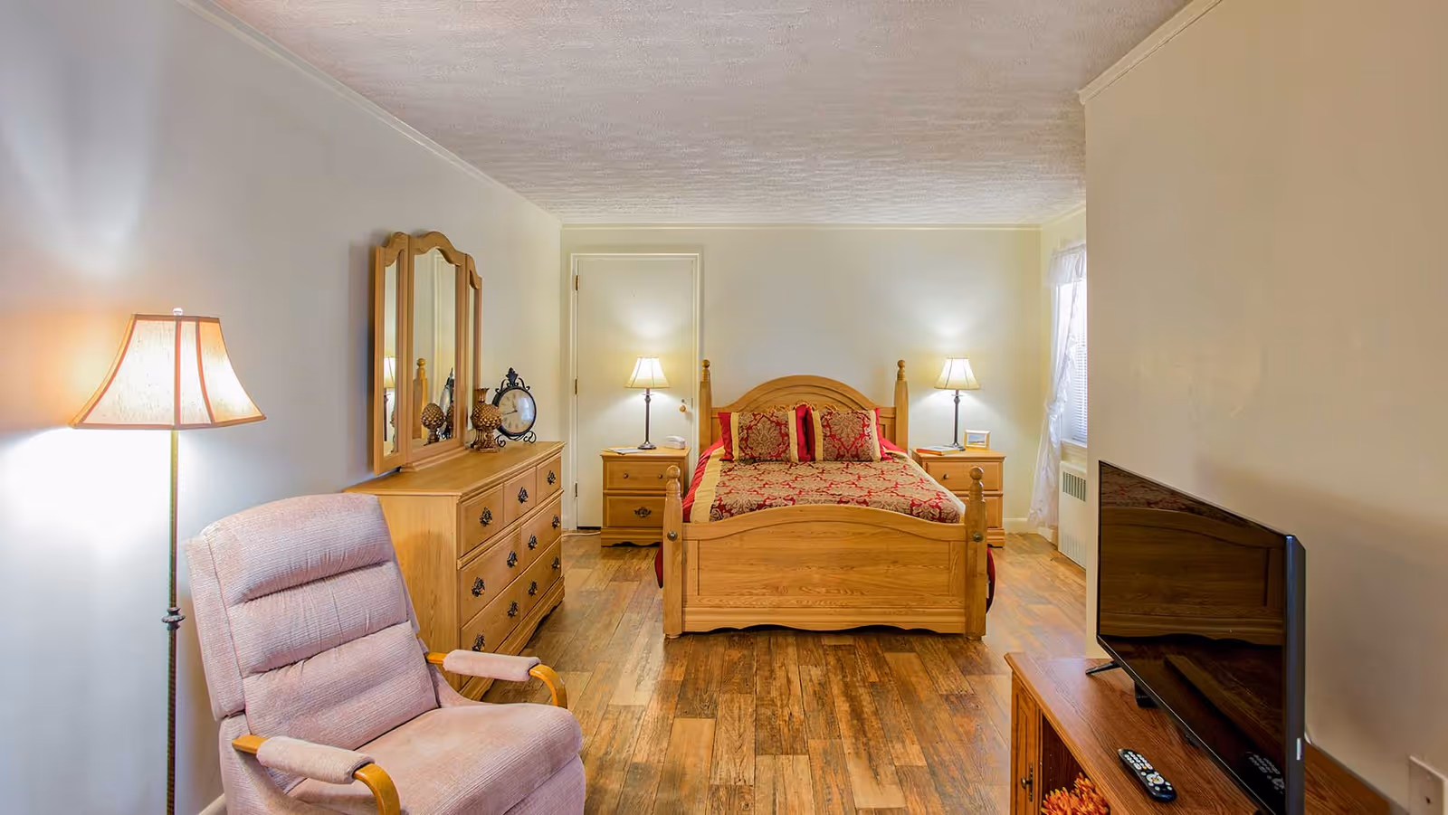 A cozy bedroom with wooden furniture including a bed with red and gold bedding, two bedside tables with lamps, a dresser with a mirror, a cushioned armchair, a floor lamp, and a flat-screen TV on a wooden stand. The room has wooden flooring and light-colored walls.