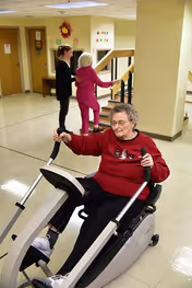 An elderly woman wearing a red sweater is using a seated exercise bike in a hallway of an assisted living facility. In the background, a younger woman is assisting another elderly woman who is holding onto a railing near a staircase. The setting appears to be indoors with beige walls and a tiled floor.