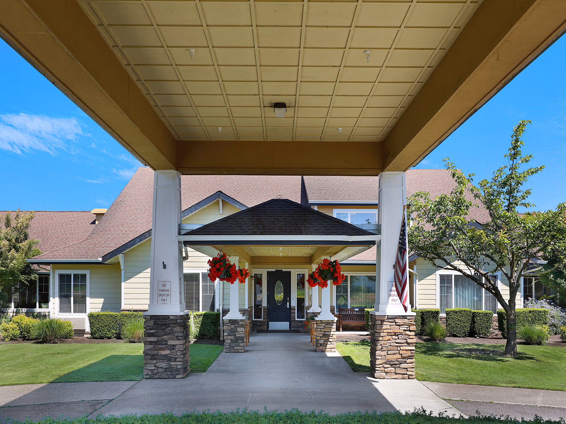 Entrance of a senior living facility with a covered driveway supported by white columns with stone bases. Hanging red flowers decorate the entrance area. The building has beige siding, large windows, and a brown roof. There is an American flag on the right side and well-maintained landscaping with green grass, bushes, and trees under a clear blue sky.