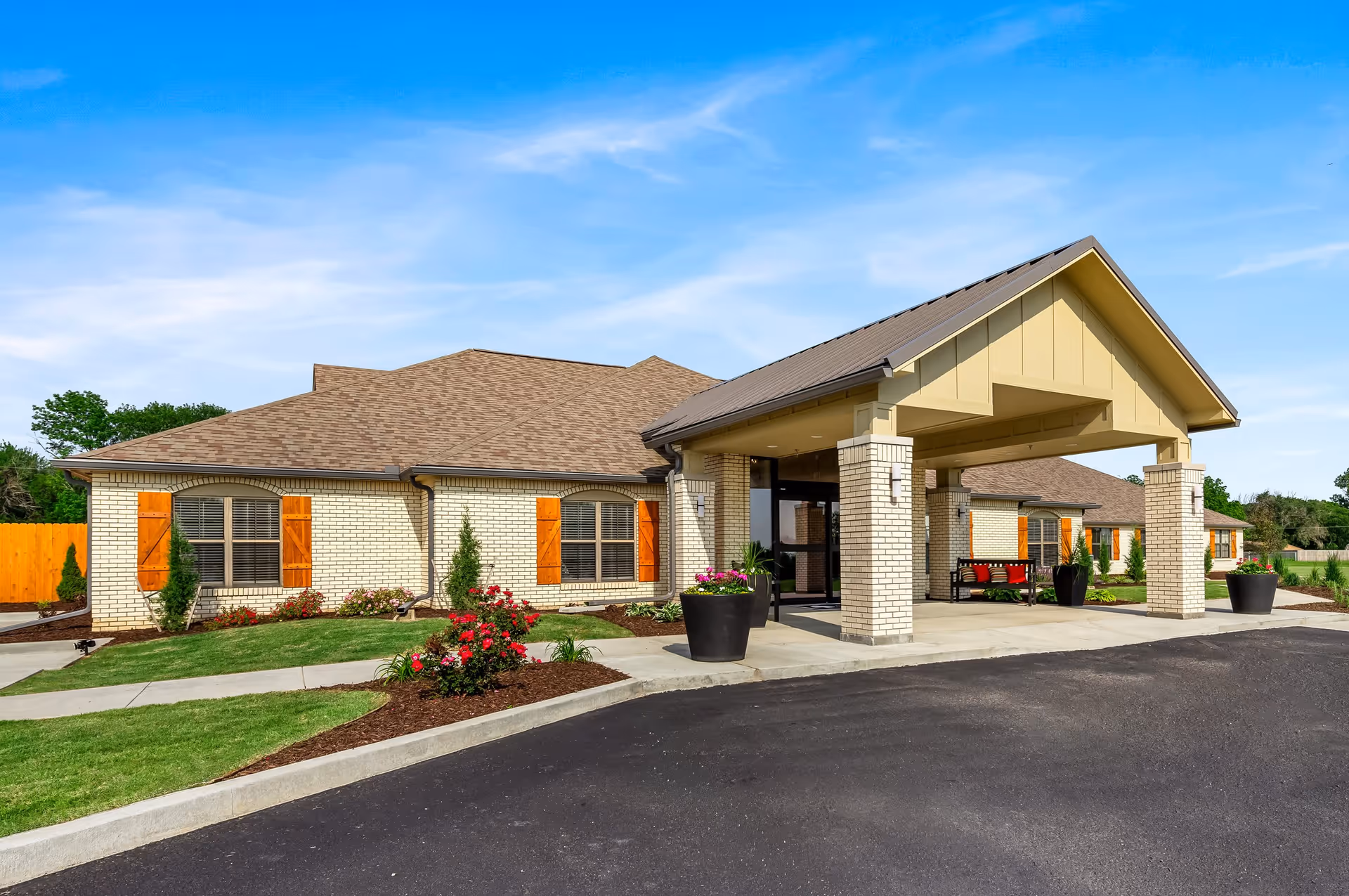 Exterior view of Asbury Village building with beige brick walls, brown shingled roof, wooden window shutters, and a covered entrance supported by brick pillars. The surrounding area includes a paved driveway, landscaped garden beds with flowers and shrubs, and a clear blue sky.