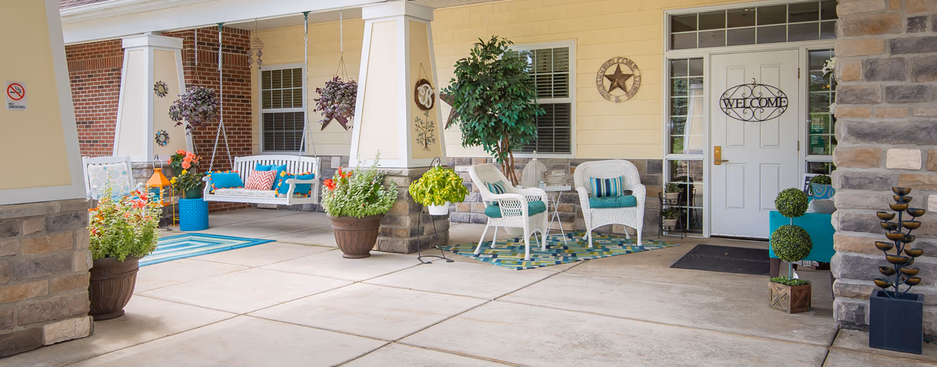 Covered outdoor seating area with white wicker chairs, a white porch swing with colorful cushions, potted plants, and decorative wall hangings including a welcome sign on a white door.