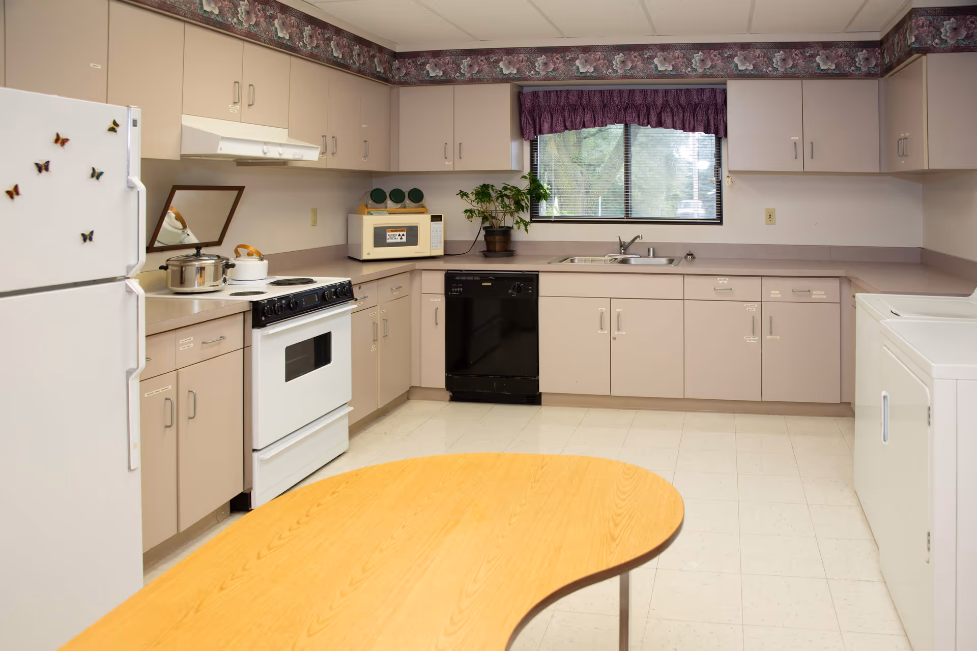 A clean kitchen with beige cabinets, a white refrigerator decorated with butterfly magnets, a white stove with pots on it, a microwave, a black dishwasher, a sink under a window with a purple valance, and a wooden table in the foreground.