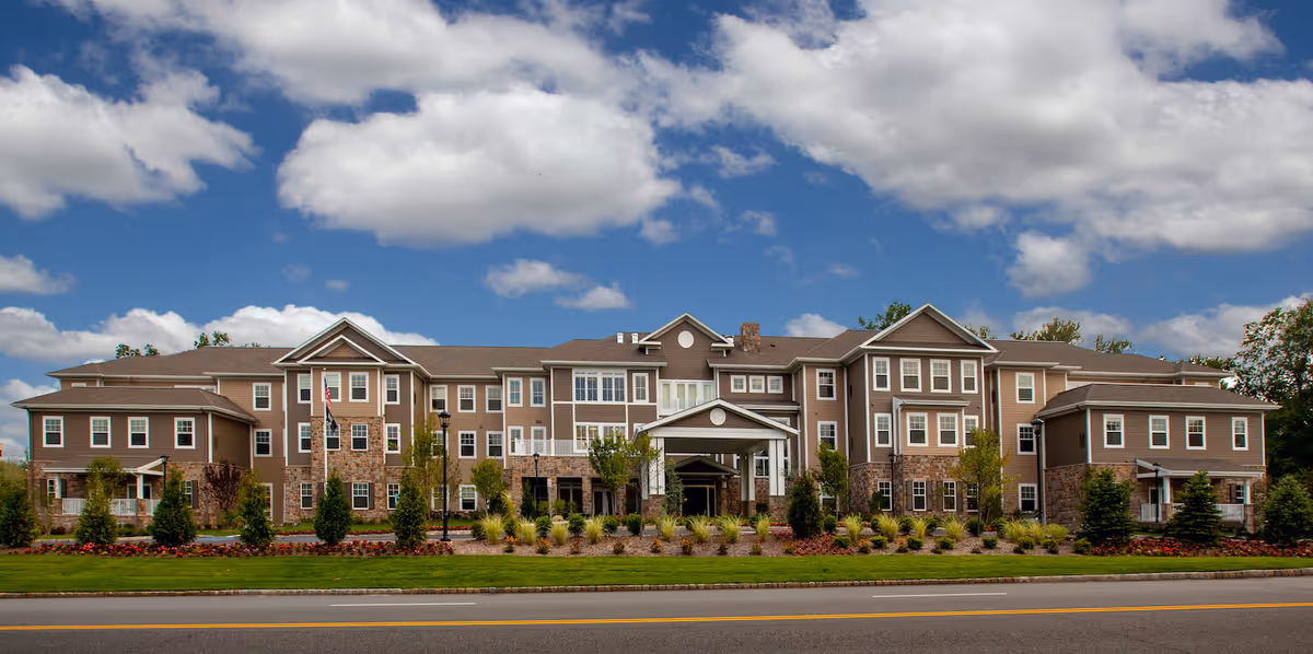 Front view of a three-story senior living building with a covered entrance and landscaped lawn under a blue sky.