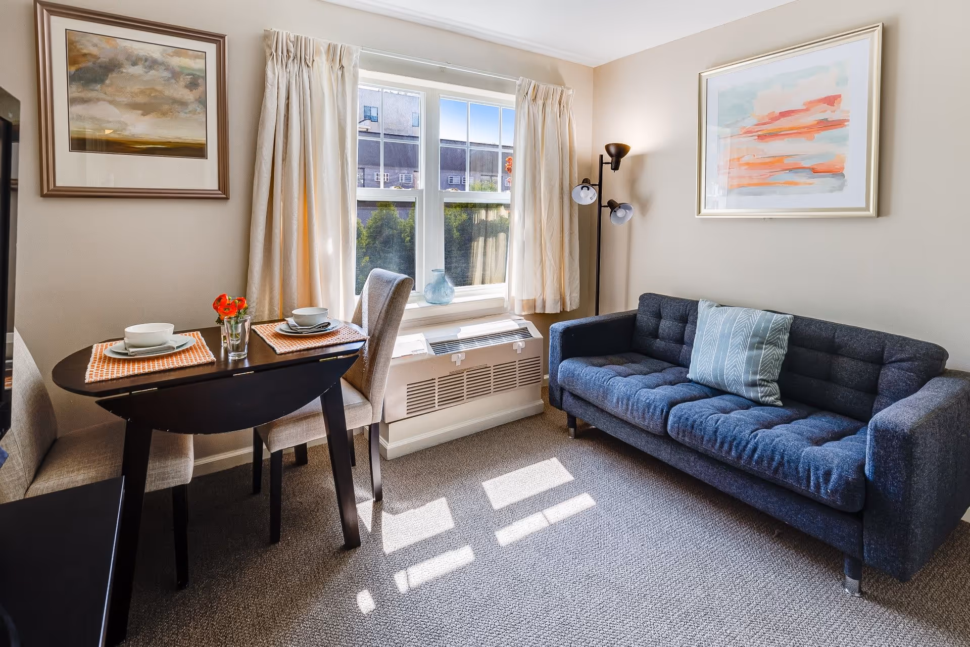 A bright living room area with a dark blue tufted sofa with a patterned pillow, a small dark wood dining table set for two with beige chairs, a window with cream curtains letting in sunlight, and two framed abstract paintings on the walls.
