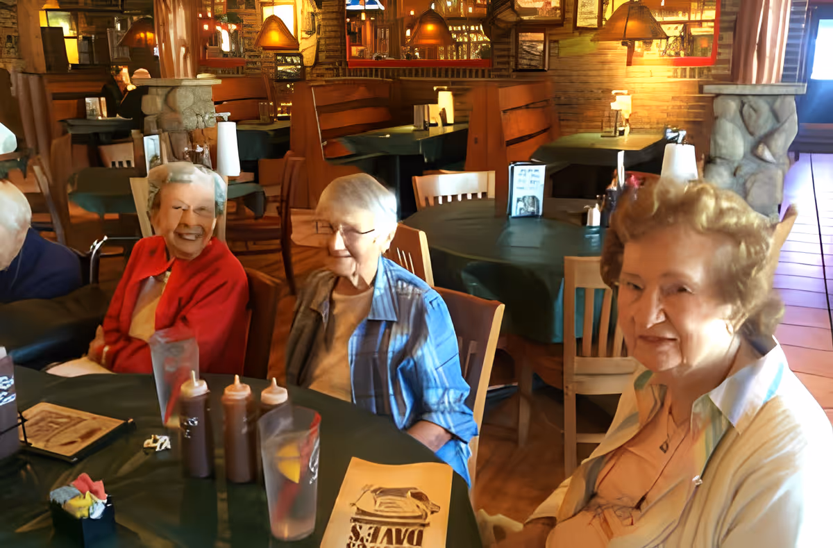 Three elderly women sitting around a table in a warmly lit restaurant or dining area with wooden furniture and stone pillars. The table has menus, glasses of water with lemon, and condiment bottles. The background shows more tables and booths with green tablecloths and hanging lamps.