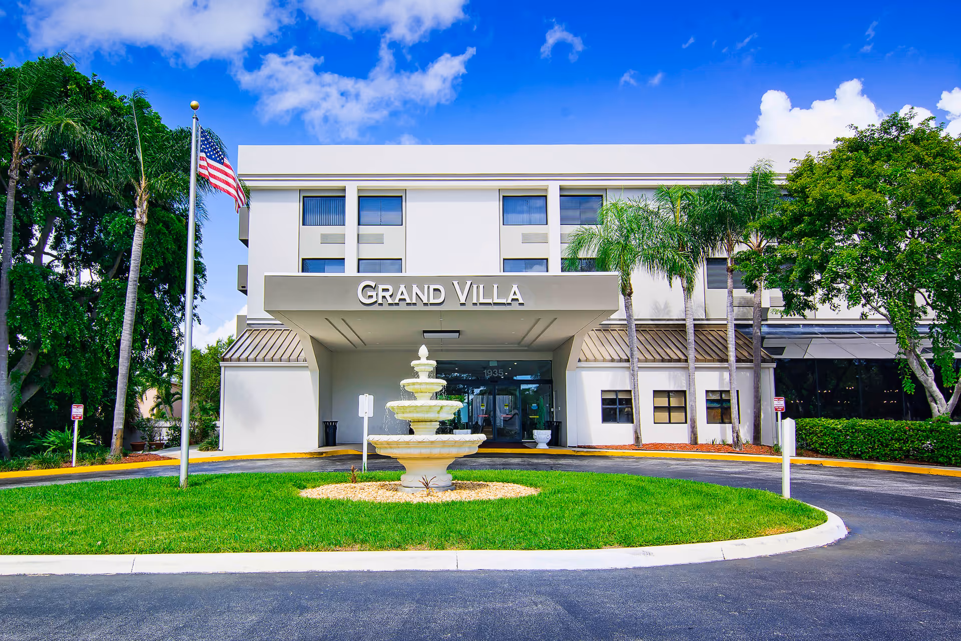 Front exterior view of the Grand Villa building with a three-tiered water fountain in the center of a circular driveway, palm trees, an American flag on a flagpole, and a bright blue sky with some clouds.