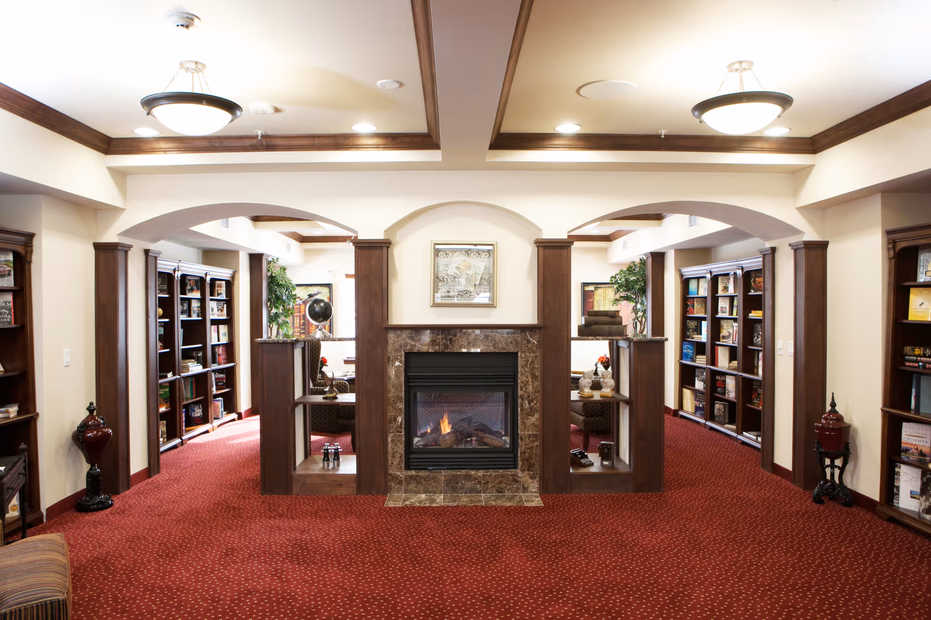 A cozy interior room with a red carpet and wooden trim featuring a central fireplace with a marble surround. On either side of the fireplace are built-in shelves with decorative items and books. The room has arched openings leading to areas with more bookshelves and seating, illuminated by ceiling lights.