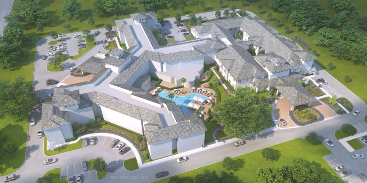 Aerial view of a large senior living facility with multiple connected buildings surrounding a central courtyard featuring a swimming pool, lounge chairs, and shaded seating areas. The facility is surrounded by parking lots, roads, and green landscaped areas with trees and grass.