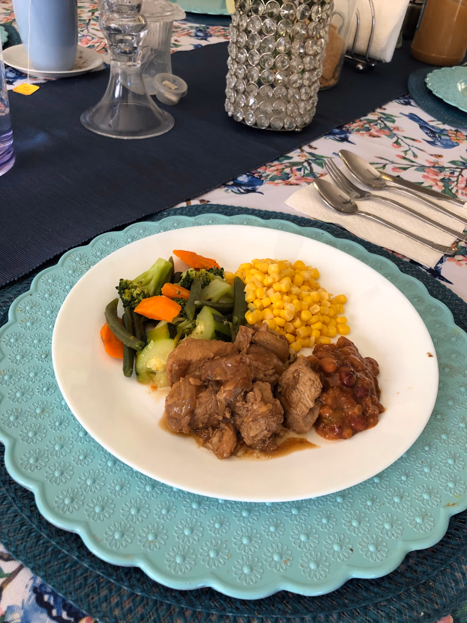 A white plate with cooked vegetables including broccoli, carrots, green beans, corn, and a serving of meat with gravy and baked beans, placed on a turquoise decorative placemat on a dining table with floral tablecloth and silverware.