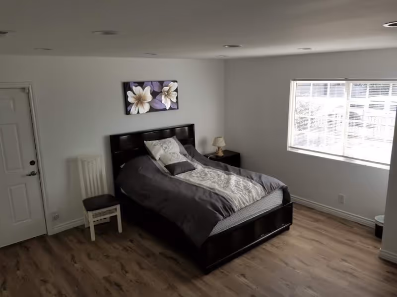 A simple bedroom with a dark wooden bed frame and a bed made with gray and white bedding. There is a white chair with a dark cushion next to the bed, a small nightstand with a lamp, a window with blinds letting in natural light, and a floral painting on the wall above the bed. The floor is wooden and the walls are white.