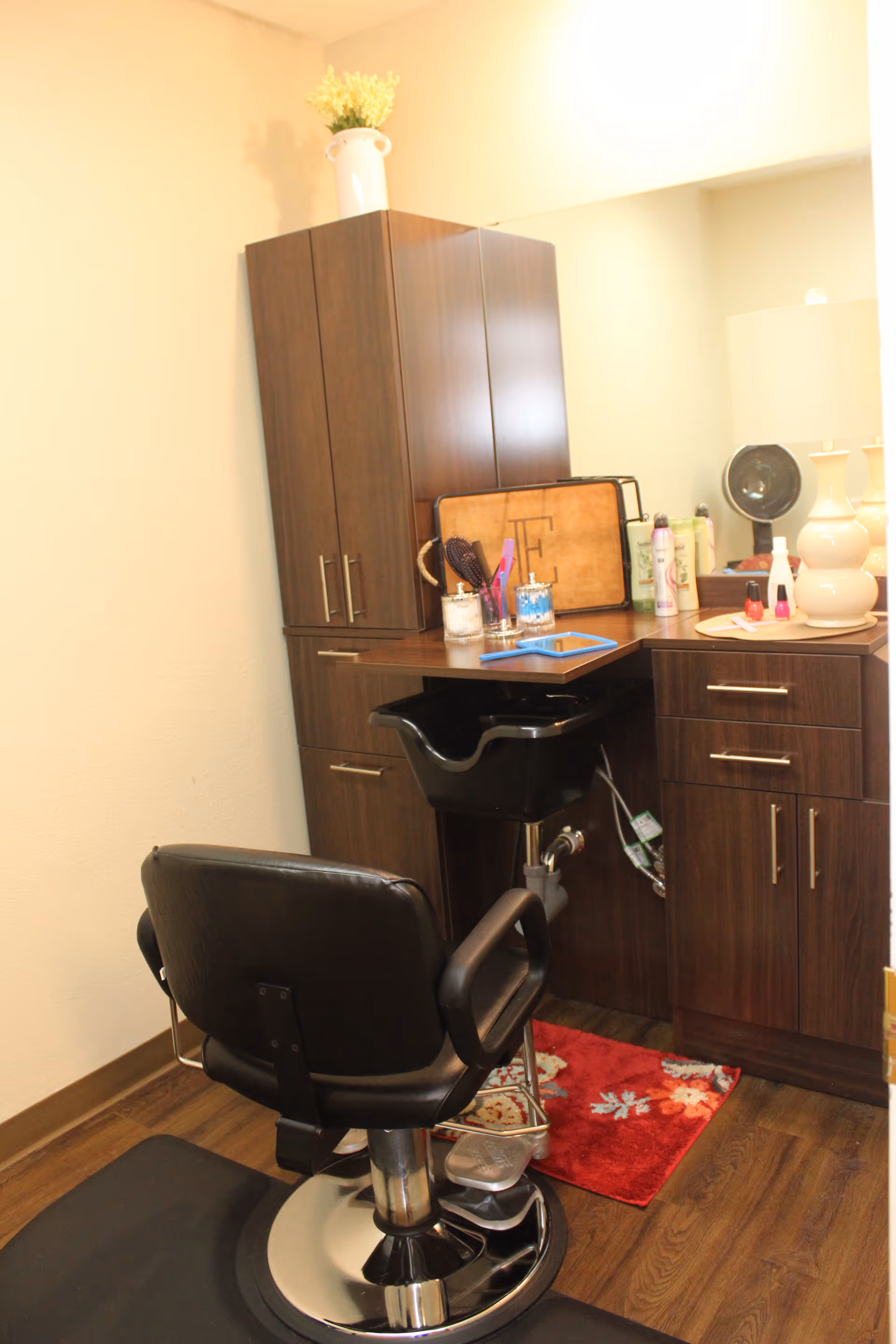 A small hair salon station inside a room with a black salon chair, a black hair washing sink, dark wood cabinets, a mirror, and various hair and beauty products on the countertop. A white lamp and a vase with flowers are also visible on the counter.