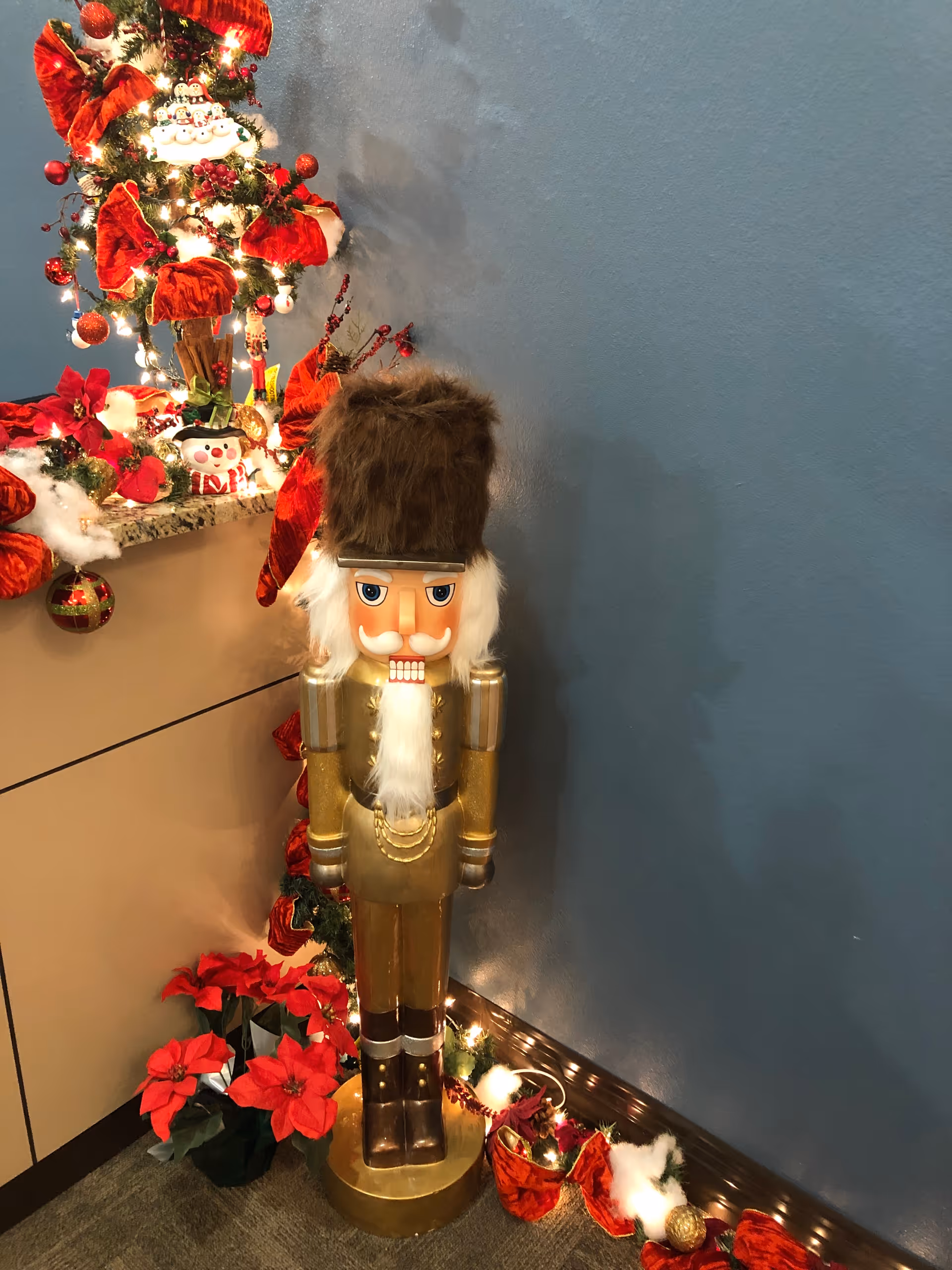 A festive holiday display featuring a large golden nutcracker figure with a furry hat standing next to a decorated corner with red poinsettias, a small Christmas tree adorned with red bows and ornaments, and string lights against a blue wall.