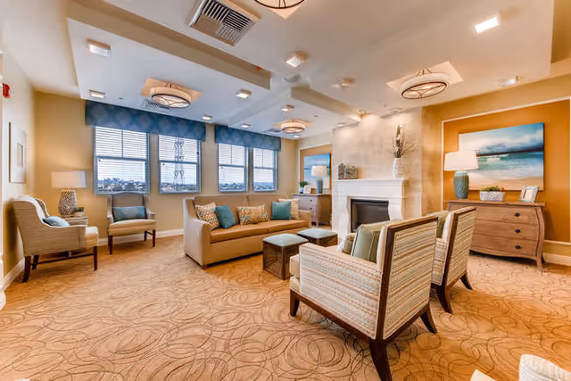 A spacious and well-lit living room with beige walls and carpet featuring a circular pattern. The room includes a beige sofa with blue and patterned cushions, two striped armchairs, a fireplace with a stone surround, a wooden dresser with a lamp and decorative items, and large windows with blue valances allowing natural light to enter.