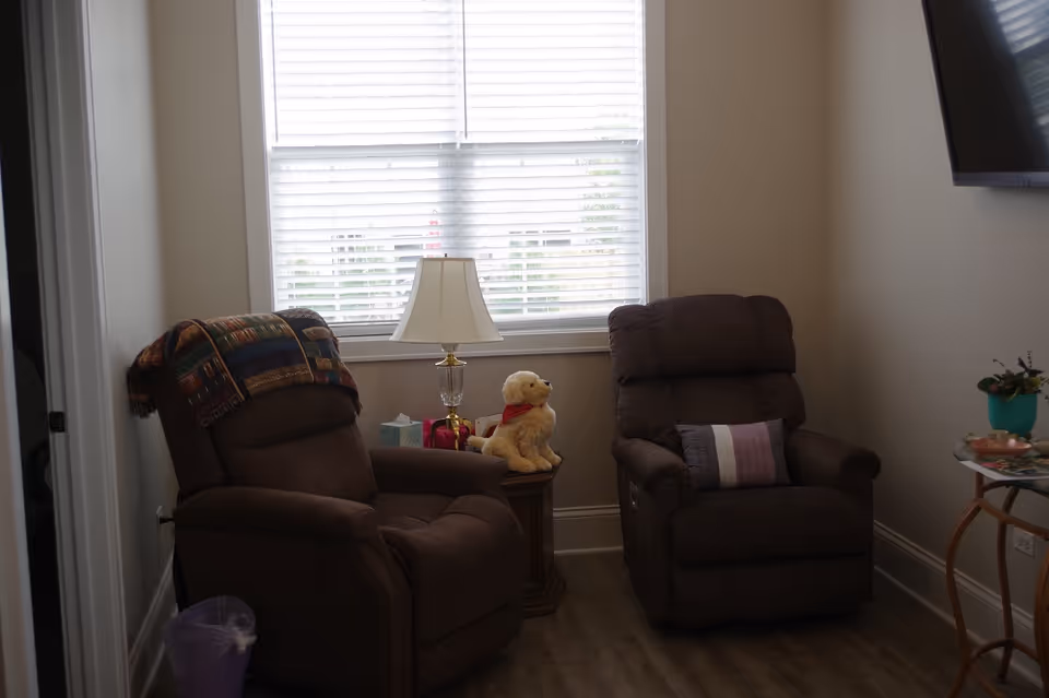 A cozy sitting area with two brown recliner chairs positioned near a window with white blinds. Between the chairs is a small wooden side table holding a lamp, a tissue box, and a stuffed dog toy wearing a red scarf. A colorful blanket is draped over one chair, and a small round glass table with a plant and some items is visible on the right side. The room has light-colored walls and wood flooring.