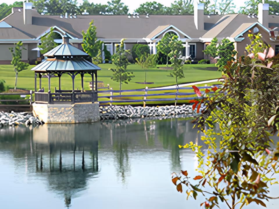 A peaceful outdoor scene at Knolls of Oxford featuring a small gazebo on a stone platform over a calm pond, with a wooden fence and well-maintained green lawn in the background. Trees and shrubs surround the area, and residential buildings are visible behind the lawn.