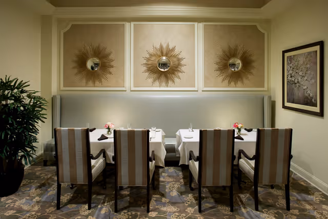 A dining area with two tables covered in white tablecloths, each set with glasses, plates, and napkins. Six striped upholstered chairs are arranged around the tables. The back wall features a cushioned bench seat and three decorative sunburst mirrors. A potted plant is on the left side and a framed floral painting hangs on the right wall.