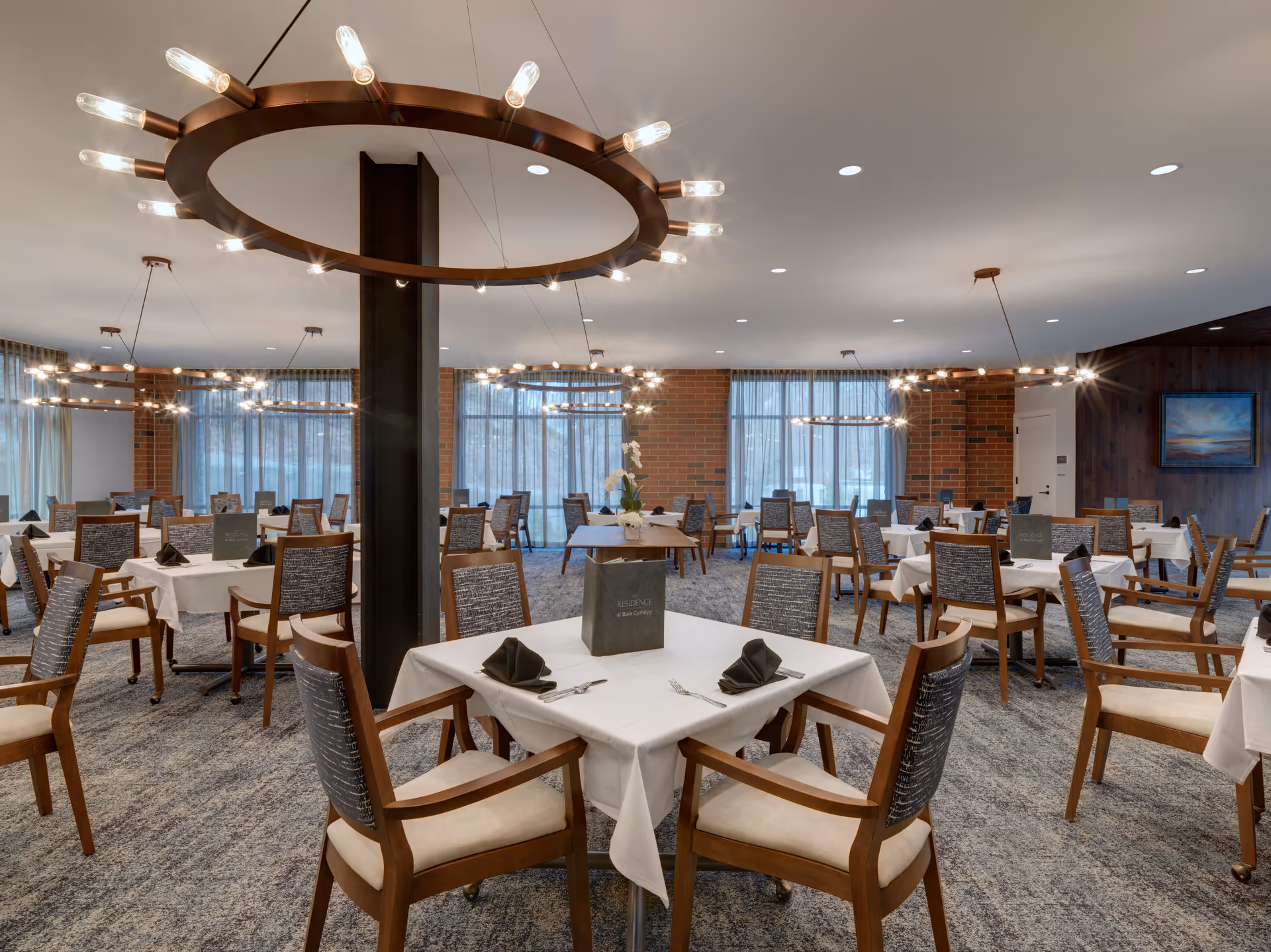 Spacious dining room with multiple tables set with white tablecloths and wooden chairs under circular chandeliers.