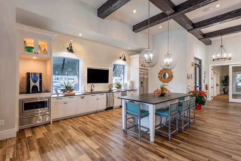 Open modern kitchen and communal dining area with a large island, pendant lights, exposed wood beams, and stainless steel appliances.