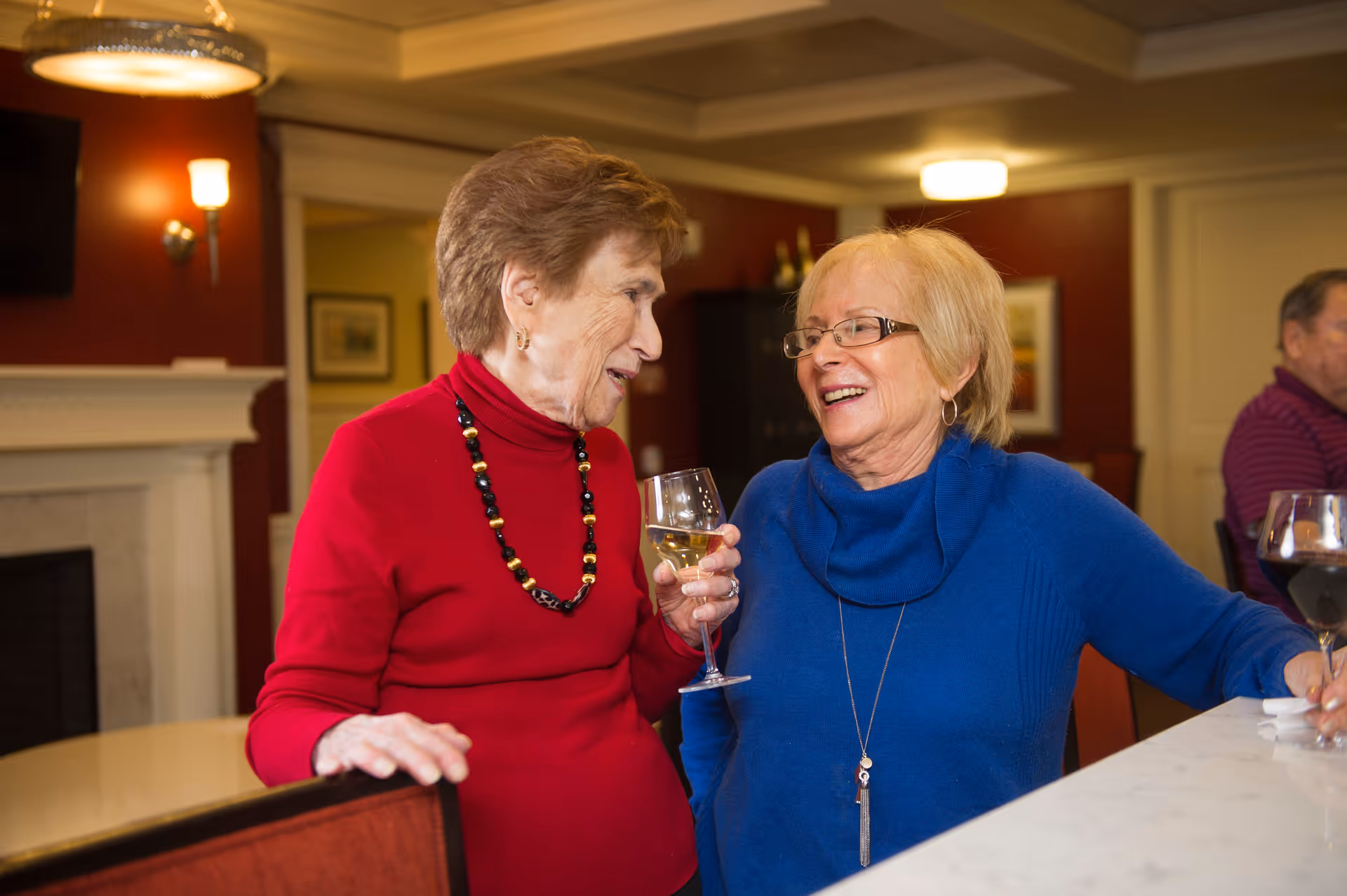 Two elderly women smiling and enjoying drinks while standing at a bar or counter in a warmly lit room with red walls and a fireplace in the background.