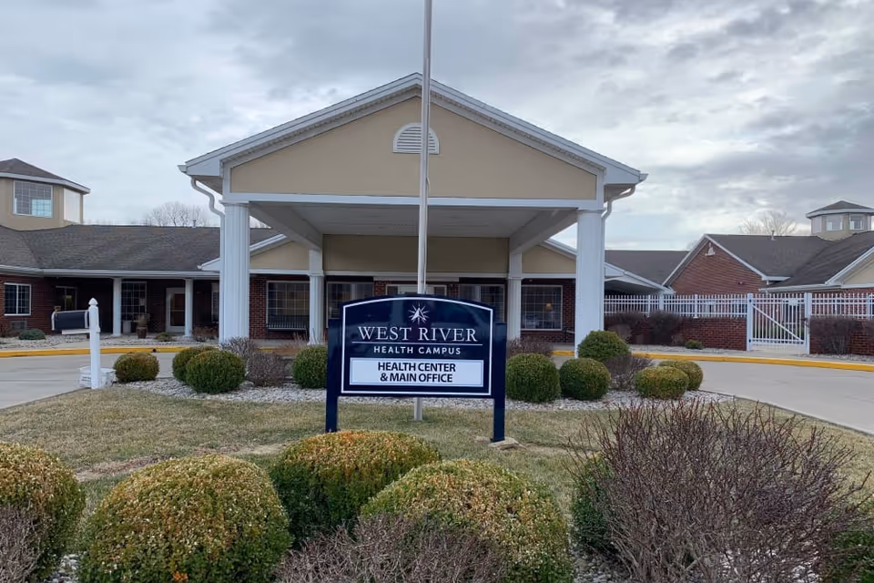 Front entrance of West River Health Campus with a sign reading 'West River Health Campus Health Center & Main Office' and landscaped bushes in front.