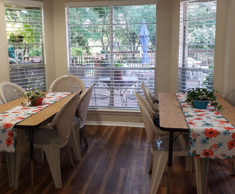 A dining area with two rectangular tables covered with floral table runners and small potted plants. Each table is surrounded by white plastic chairs. Large windows with blinds reveal an outdoor patio with more tables, chairs, and greenery.