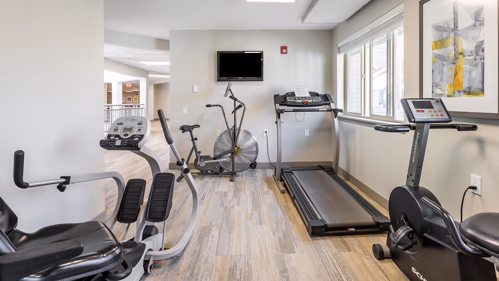 A small fitness room with exercise equipment including a recumbent bike, an upright stationary bike, a treadmill, and an air resistance exercise bike. The room has light-colored walls and flooring, a large window letting in natural light, a wall-mounted TV, and a modern abstract painting on the wall.