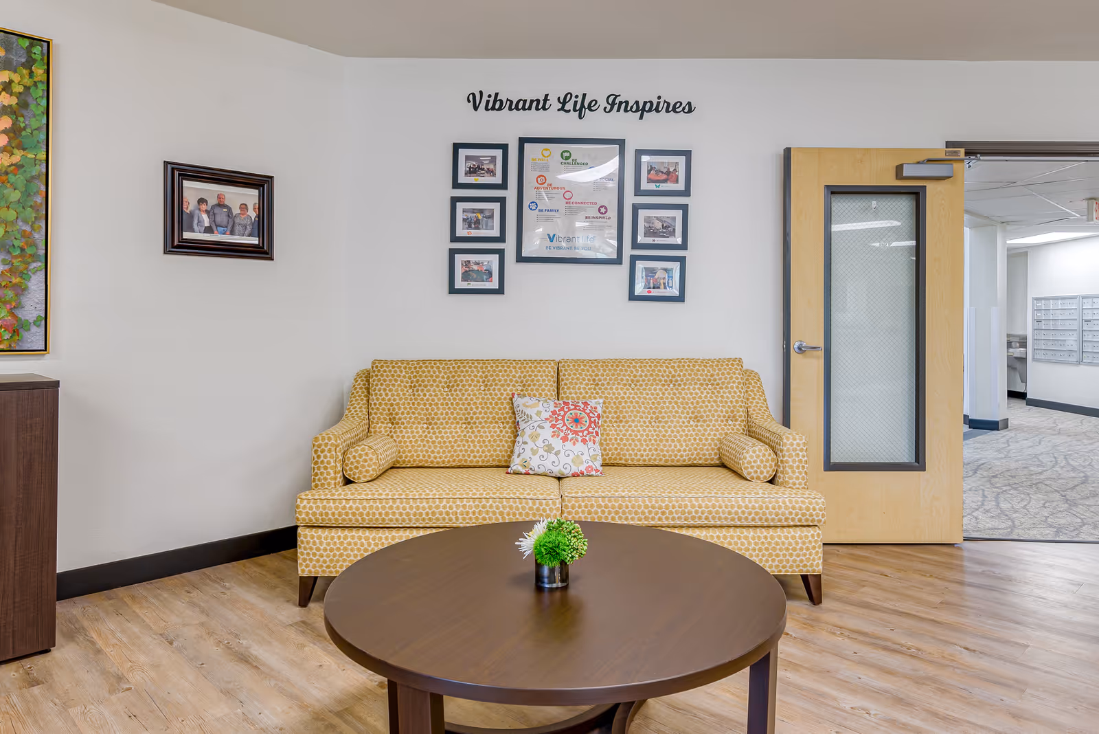 Small seating area with a yellow patterned sofa, round wooden coffee table and framed photos beneath the words "Vibrant Life Inspires" on a white wall.