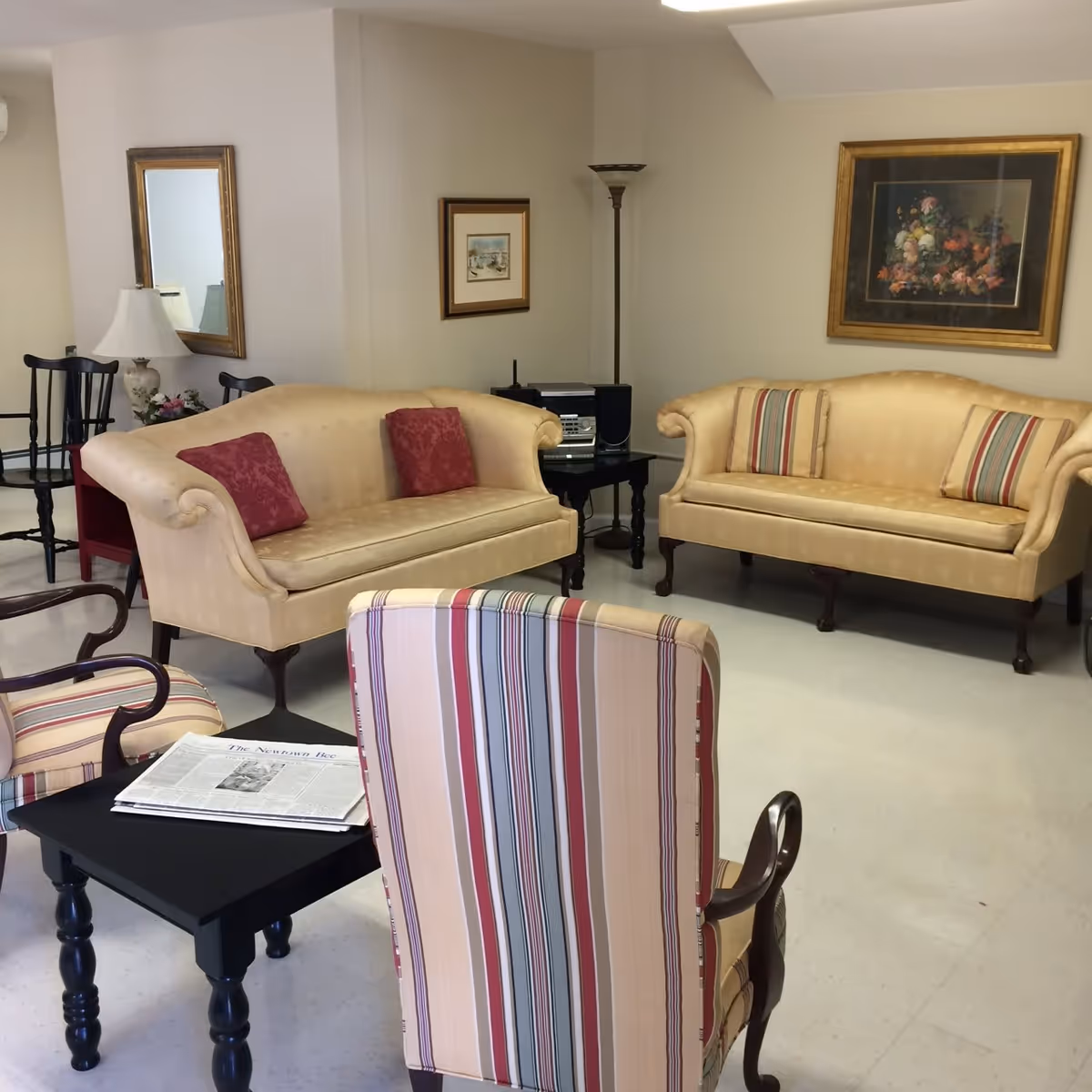 A sitting room with two beige sofas, striped armchairs, a black coffee table with a newspaper, and framed wall art on the walls.