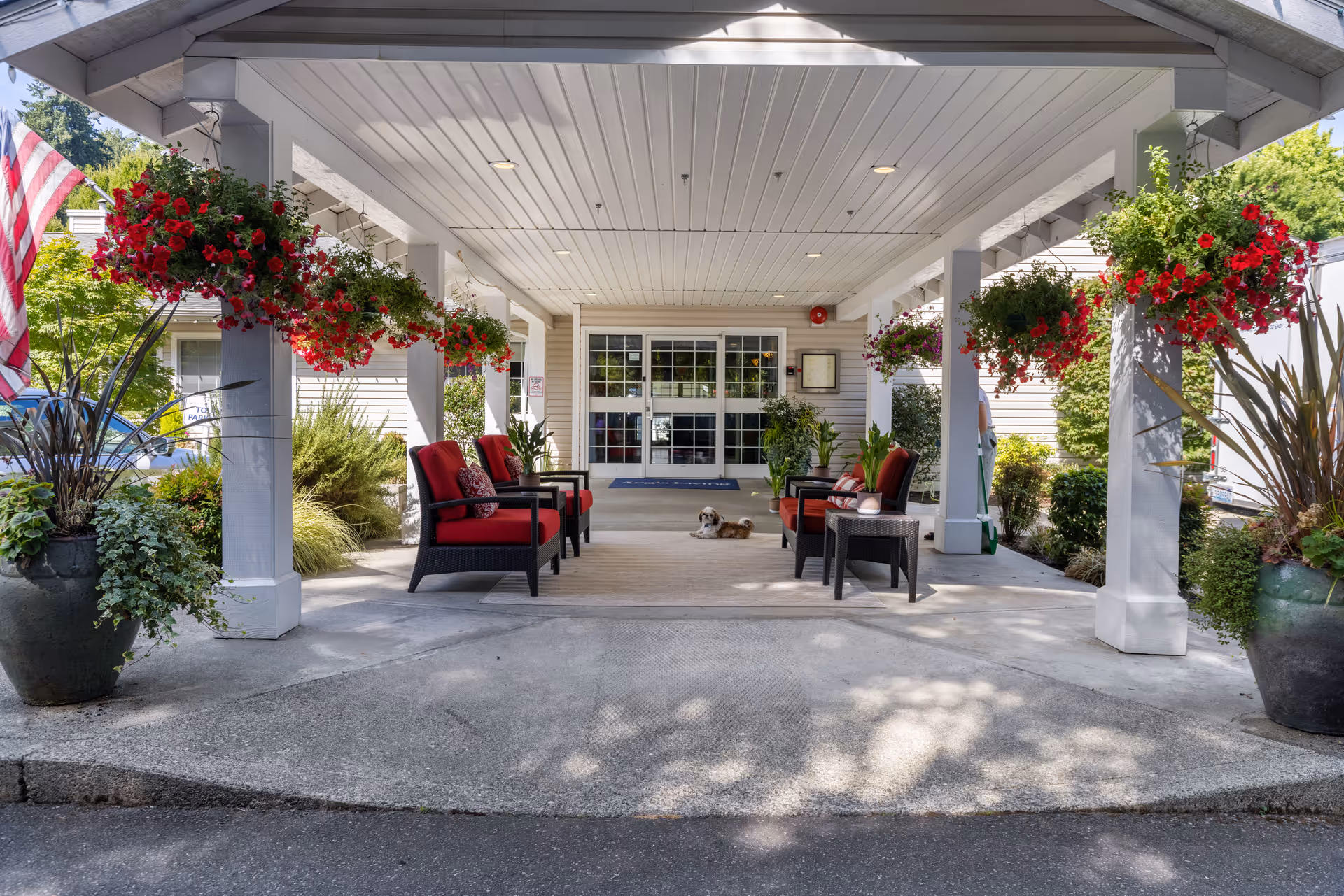 Covered entrance area of a senior living facility with red cushioned chairs arranged on a rug, hanging flower baskets with red flowers, large potted plants, and an American flag on the left. A small dog is lying on the rug near the entrance doors.