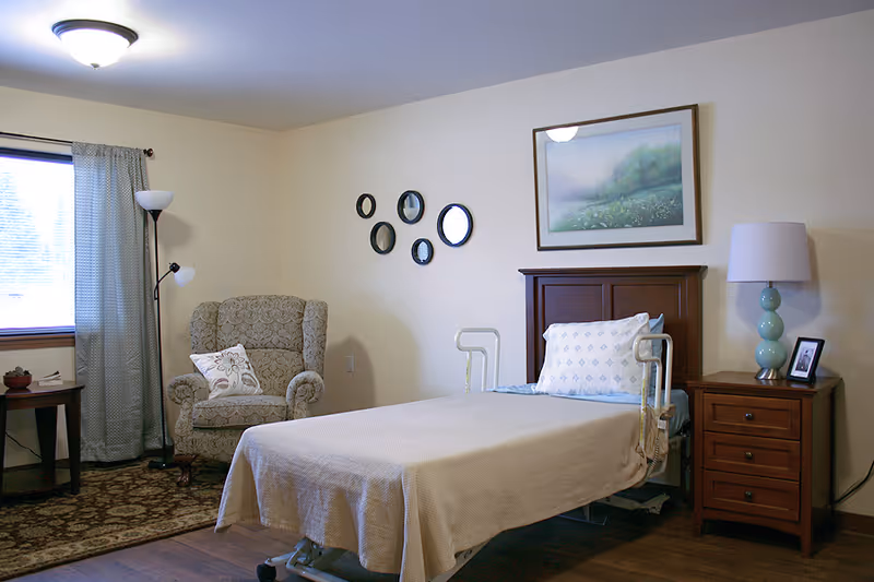 Furnished assisted living bedroom with a single adjustable bed, wooden headboard and nightstand, an armchair, lamps and wall art.
