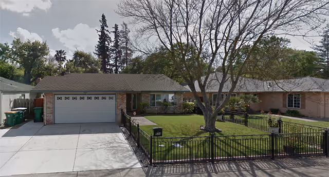 Front exterior view of a single-story residential building with a two-car garage, a driveway, a fenced front yard with a large tree, and some bushes. The sky is partly cloudy and there are tall trees in the background.