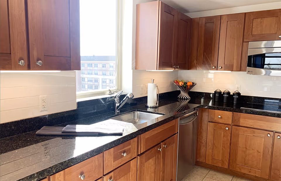 A bright kitchen with wooden cabinets, black granite countertops, a stainless steel sink, and a window letting in natural light. There is a paper towel holder, a fruit basket with assorted fruits, and three black canisters labeled tea, coffee, and sugar on the counter.