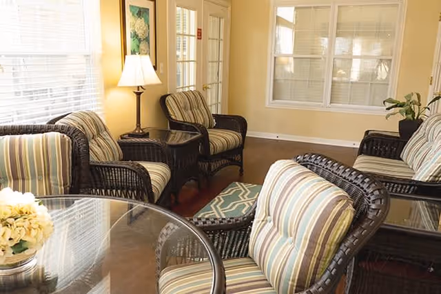 A cozy seating area with wicker chairs featuring striped cushions arranged around a glass-top table. A lamp on a side table and large windows with blinds provide natural light to the room.