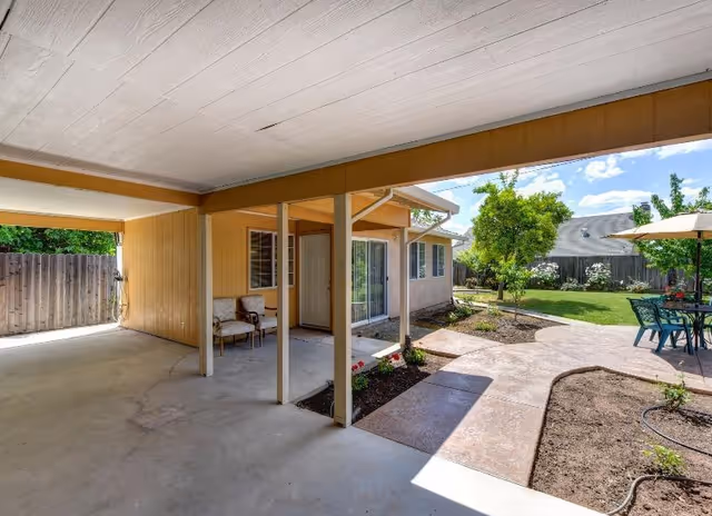 Covered patio area with concrete flooring and two chairs, adjacent to a house with beige siding and a white door. The patio opens to a landscaped backyard with a curved concrete walkway, green grass, trees, and an outdoor table with chairs under a large umbrella. The sky is blue with some clouds.